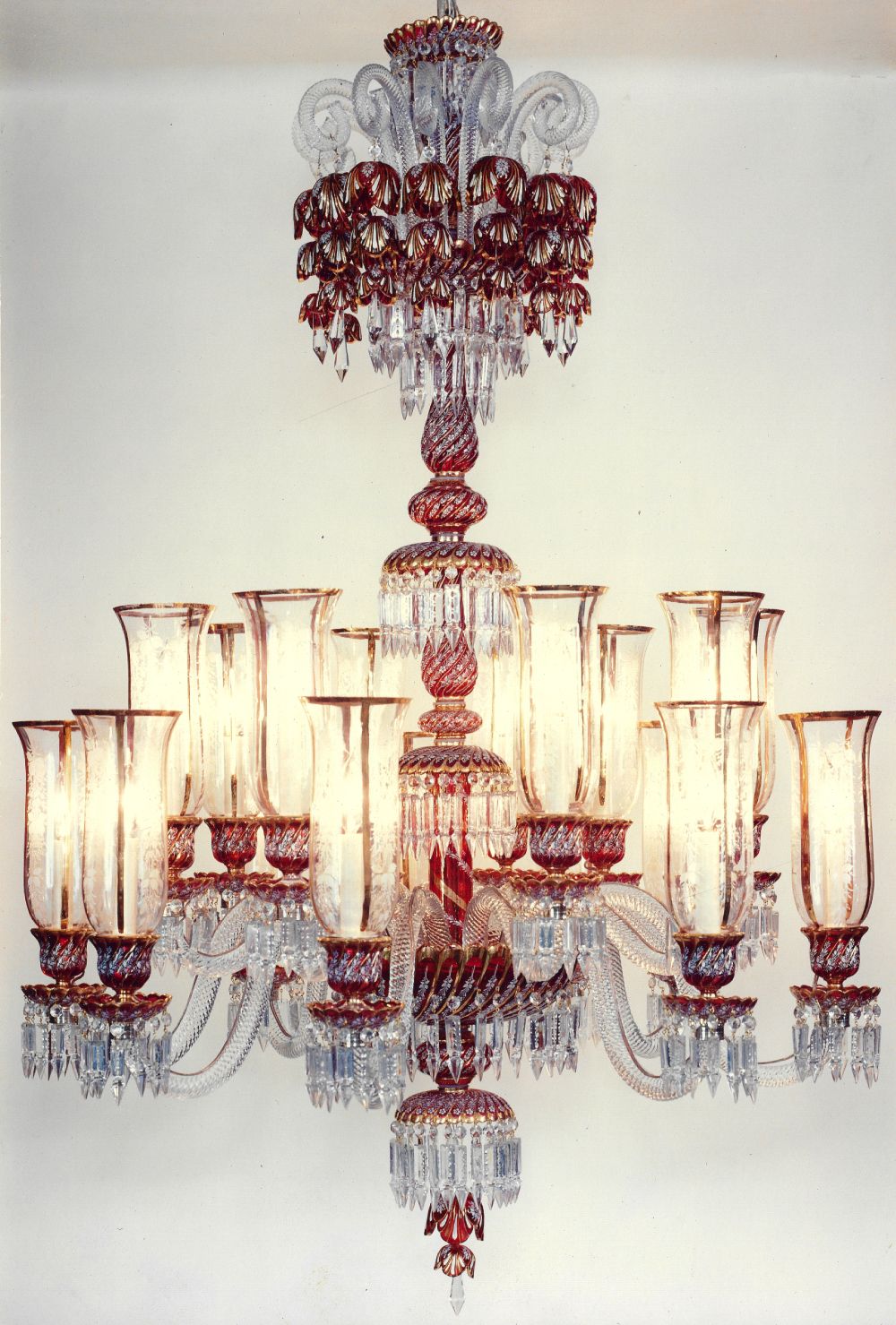 A twelve arm chandelier lit against a pale wall: ruby red glass cups and petal canopies with gilt edges, clear glass hurricane shades over each candle, spiral arms and hanging crystal prisms.