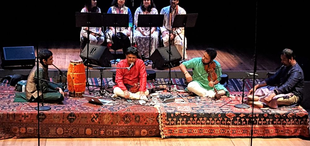 A Pakistani classical ensemble seated cross legged on layered rugs at the front of a concert stage, with a violinist, a singer, a drummer and a chorus seated behind.