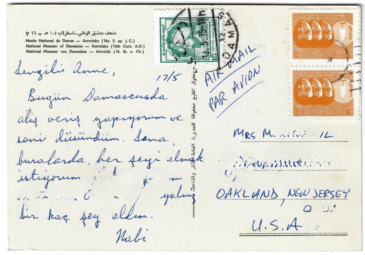 The back of a postcard, handwritten in blue ink in Turkish and signed Nabi, with two Syrian Arab Republic stamps, a Damascus postmark dated 13 May 1975, the words air mail and par avion written by hand, and an address in Oakland, New Jersey, USA.