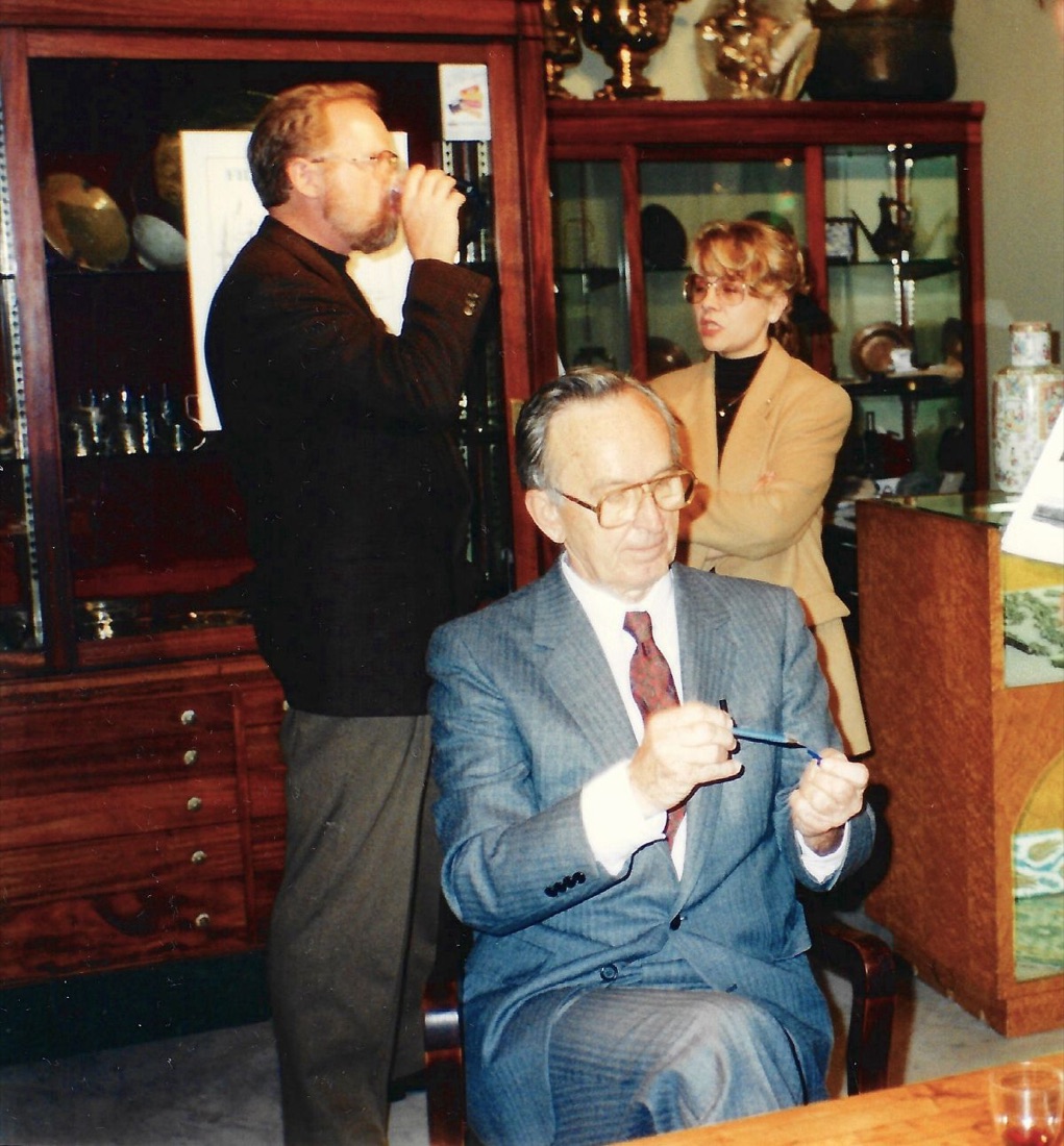 In the gallery after a lecture: an older man in a grey suit and glasses seated at a table signing books, another man standing behind him with a drink, and a woman in a tan jacket watching, with lit vitrines of metalwork behind them.