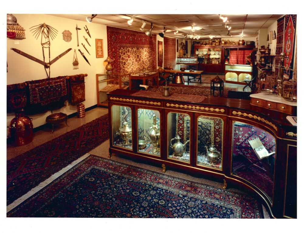 A wide view of the gallery: track lighting overhead, carpets hung on every wall, brass samovars ranked on shelves, arms and armour mounted above a bench, and a long curved vitrine of gleaming metalwork.
