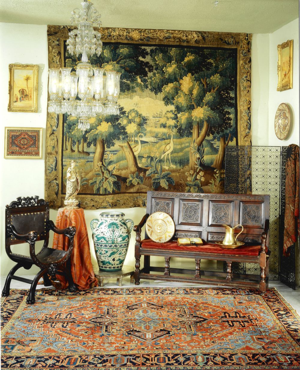 A corner of the gallery: a large verdure tapestry of trees and herons on the wall, a Venetian glass chandelier, a carved oak bench with a red cushion, a majolica jar, and a Heriz carpet on the floor.
