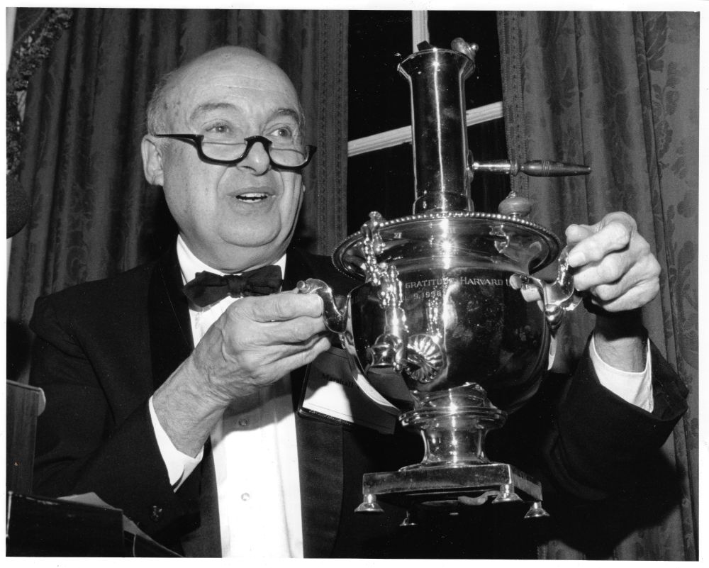 A black and white photograph: a man in a dinner jacket and bow tie holding up a large silver samovar by both handles, its body engraved with words of gratitude to Harvard.