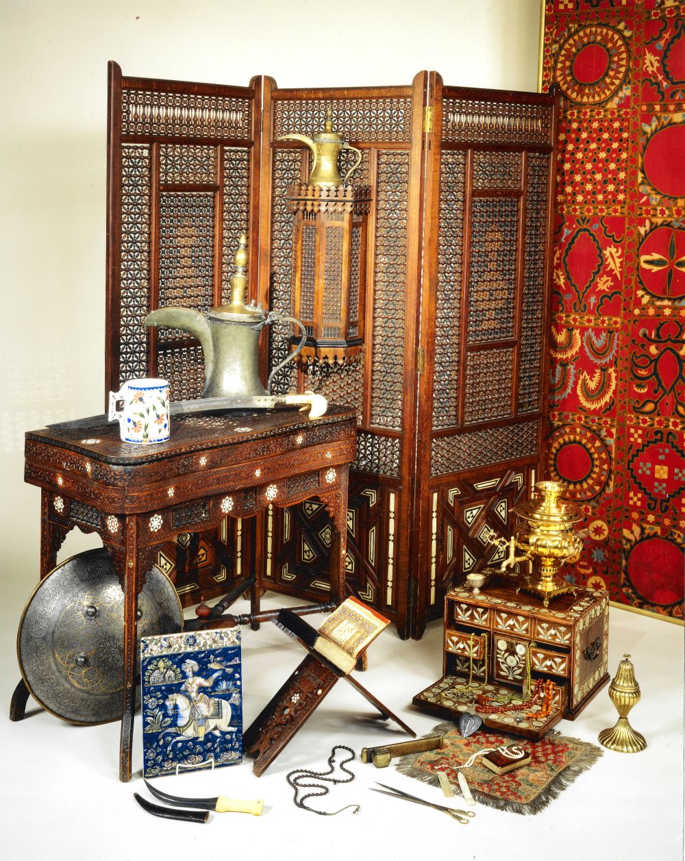 A studio photograph of Islamic works of art: a mother of pearl inlaid folding screen and card table, a large brass ewer, a damascened shield, a Persian tile of a rider on an elephant, a carved Qur'an stand, coral prayer beads, a gilt samovar and an inlaid casket.
