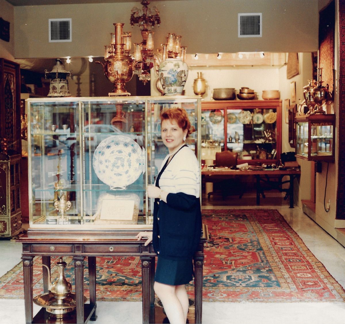A woman standing beside a tall glass vitrine in a gallery, one hand resting on the table beneath it. Inside the case a large blue and white dish on a stand; behind her brass samovars, a Chinese vase, a crystal chandelier, and a red Heriz carpet on the floor running back to the street window.