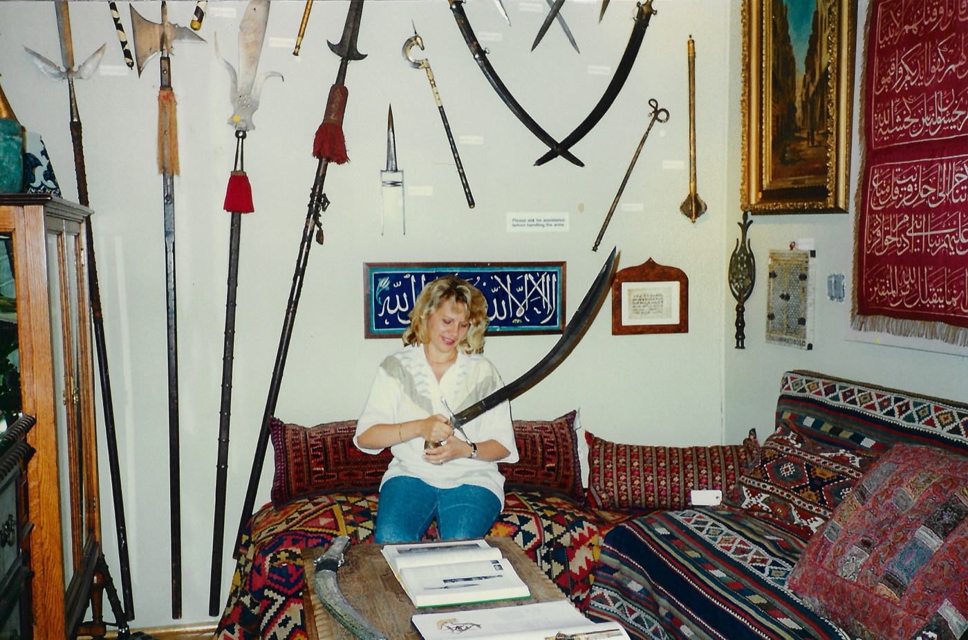 A woman seated on a kilim covered divan in a gallery, drawing a curved Ottoman sword from its scabbard and looking down at the blade. Behind her a wall hung with halberds, spears, maces, daggers and a large curved sabre, an Iznik calligraphy tile panel, a framed manuscript leaf and an embroidered red Ottoman textile.