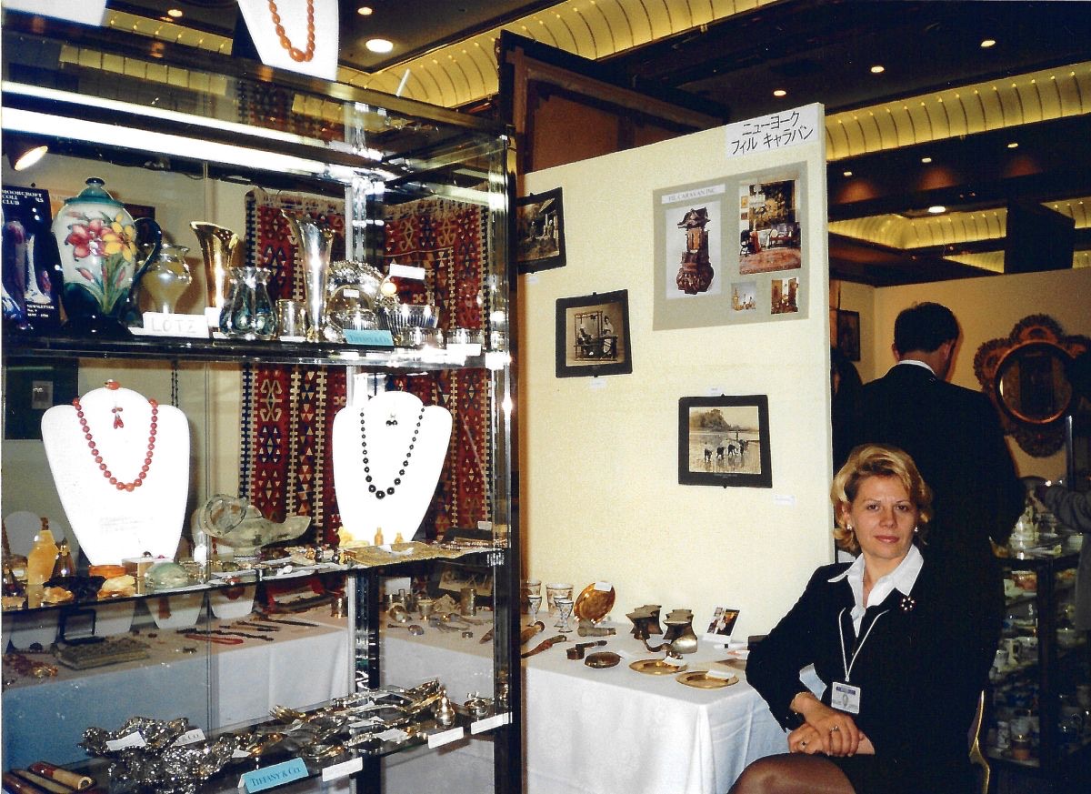 A woman seated at a trade fair stand beside a lit glass cabinet of amber and coral necklaces, silver and glass, with kilims hung behind and a panel of gallery photographs above a sign printed in Japanese katakana reading New York, Fil Caravan.