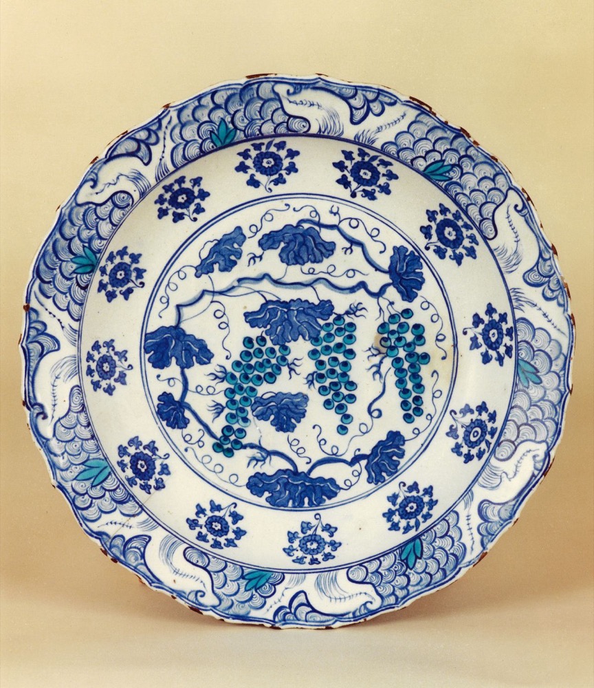 An Iznik dish painted in cobalt and turquoise: three bunches of grapes with vine leaves and tendrils in the centre, a ring of rosettes around it, and a wave and rock border on the fluted rim.
