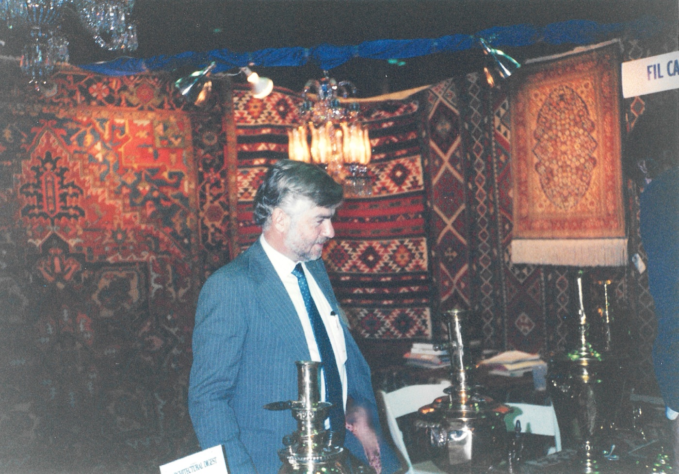 A man in a blue pinstripe suit standing in a trade fair stand hung with Caucasian rugs and striped kilims, a crystal chandelier lit above him, brass samovars and candlesticks on the table in front, and a sign reading FIL CA at the right.