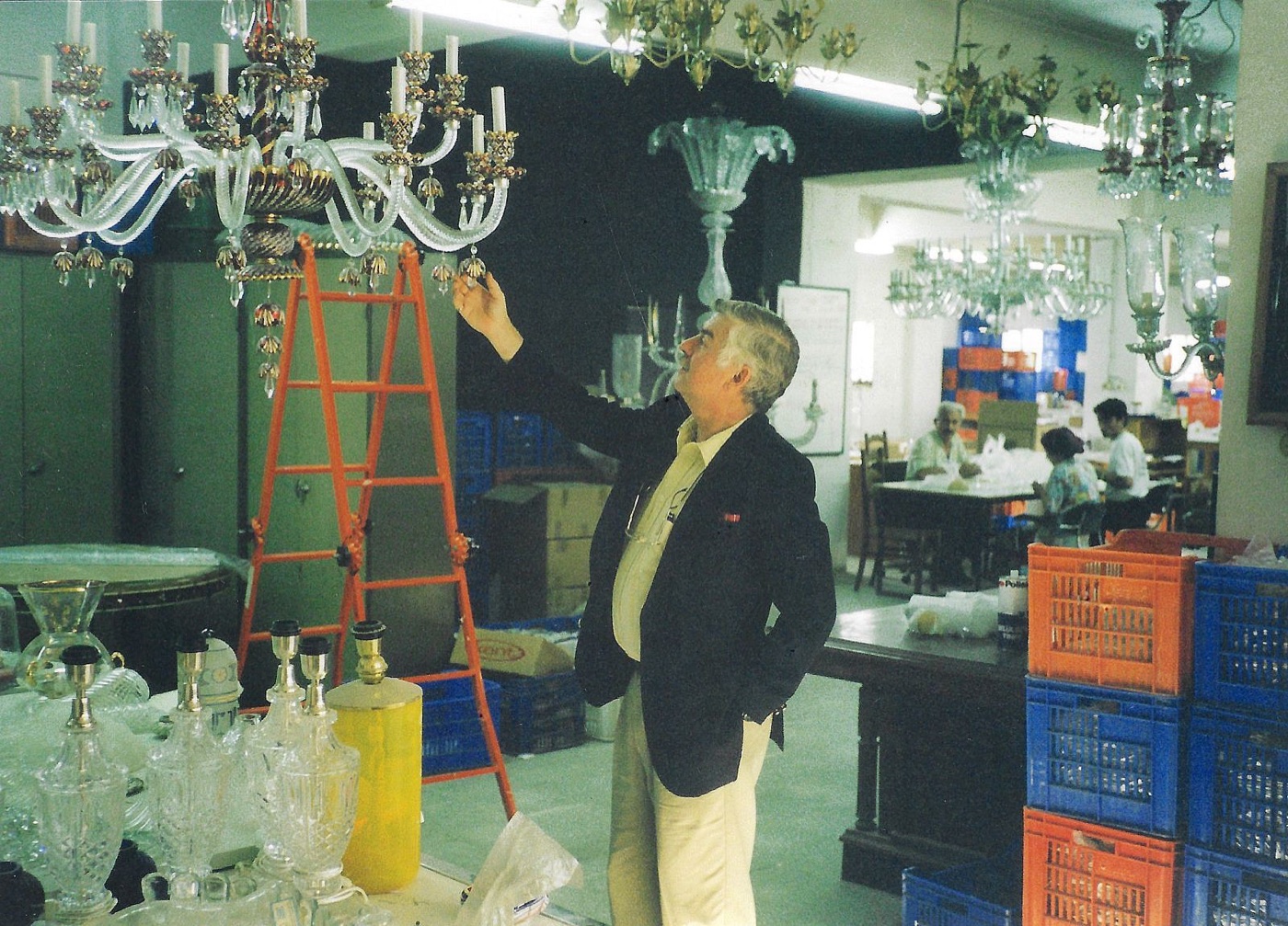 A man in a dark blazer reaching up to examine a Venetian style glass chandelier in a warehouse hung with dozens of chandeliers, with crates and a stepladder around him.