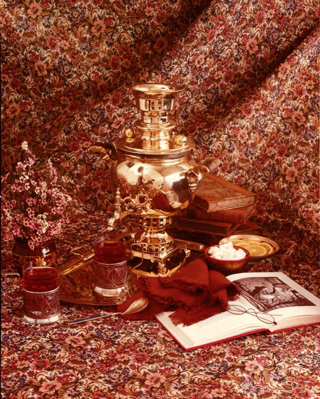 A studio still life on floral chintz: a polished brass Russian samovar with a tray, two glasses of tea in filigree holders, a bowl of sugar cubes, a dish of lemon slices, a red fringed shawl, sprays of pink blossom and an open book with spectacles resting on it.