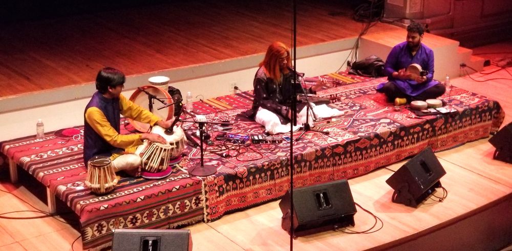 Three musicians seated on layered red and black kilims on a concert stage, one at the tabla, one at a microphone in the centre, one with a hand drum at the right.