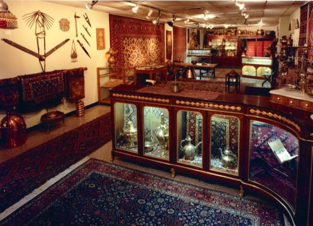 A wide view of the gallery: carpets on every wall, brass samovars on the shelves, and a long curved vitrine of metalwork.