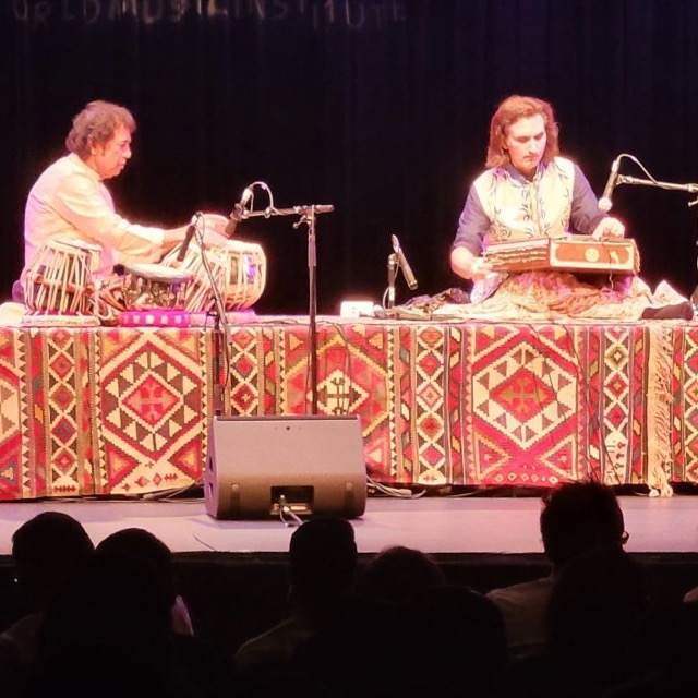 Two musicians on a concert stage, one at the tabla and one at the santoor, on a platform faced with kilims under a World Music Institute banner.