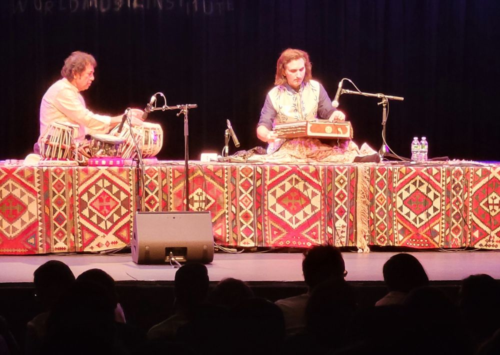 Two musicians on a lit concert stage at Symphony Space, one seated at the tabla on the left, one playing a santoor on the right, on a low platform faced with red, cream and black kilims, the words World Music Institute painted on the backdrop above them, and the heads of the audience in the dark foreground.