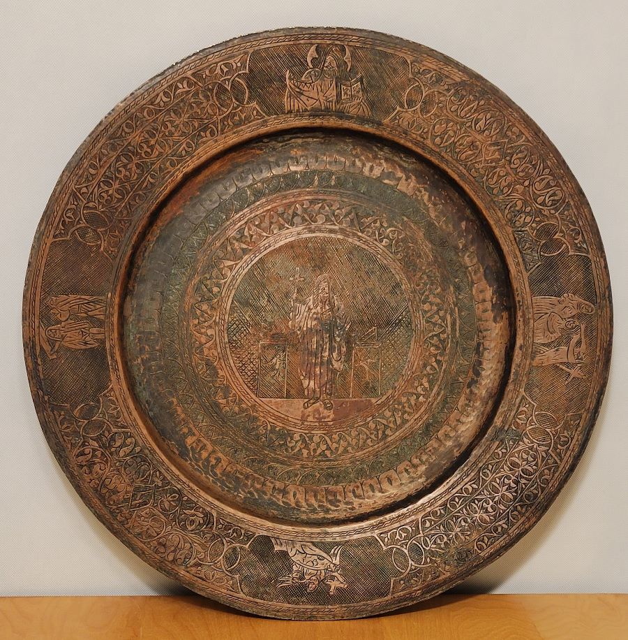 Round engraved copper alms dish with a robed saint at its center, ringed by bands of foliate ornament